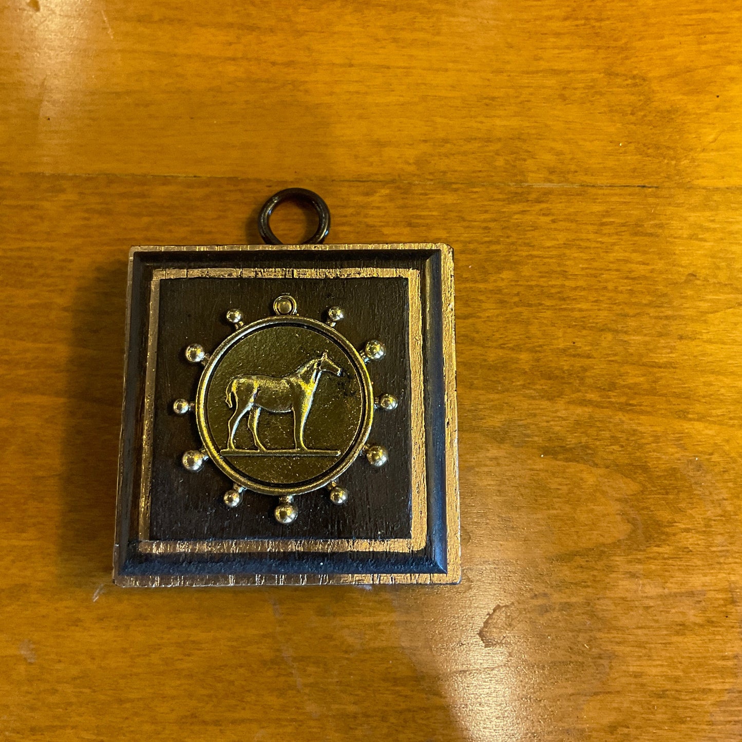 A square black and gold pendant or medallion featuring a central gold horse emblem, displayed on a warm wooden tabletop.
