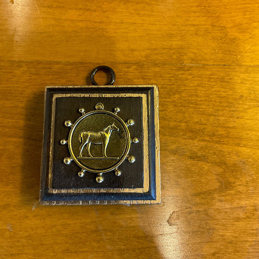 A square black and gold pendant or medallion featuring a central gold horse emblem, displayed on a warm wooden tabletop.