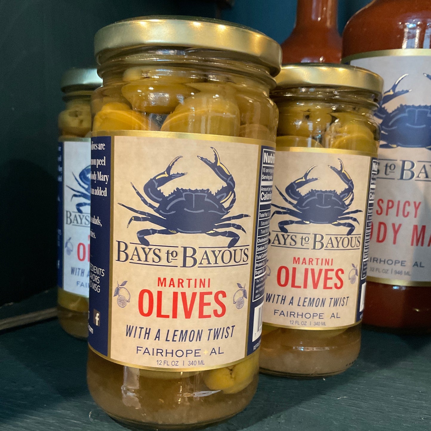 A jar of Bays to Bayous Martini Olives with a lemon twist, displayed on a dark teal shelf alongside other Bays to Bayous products.