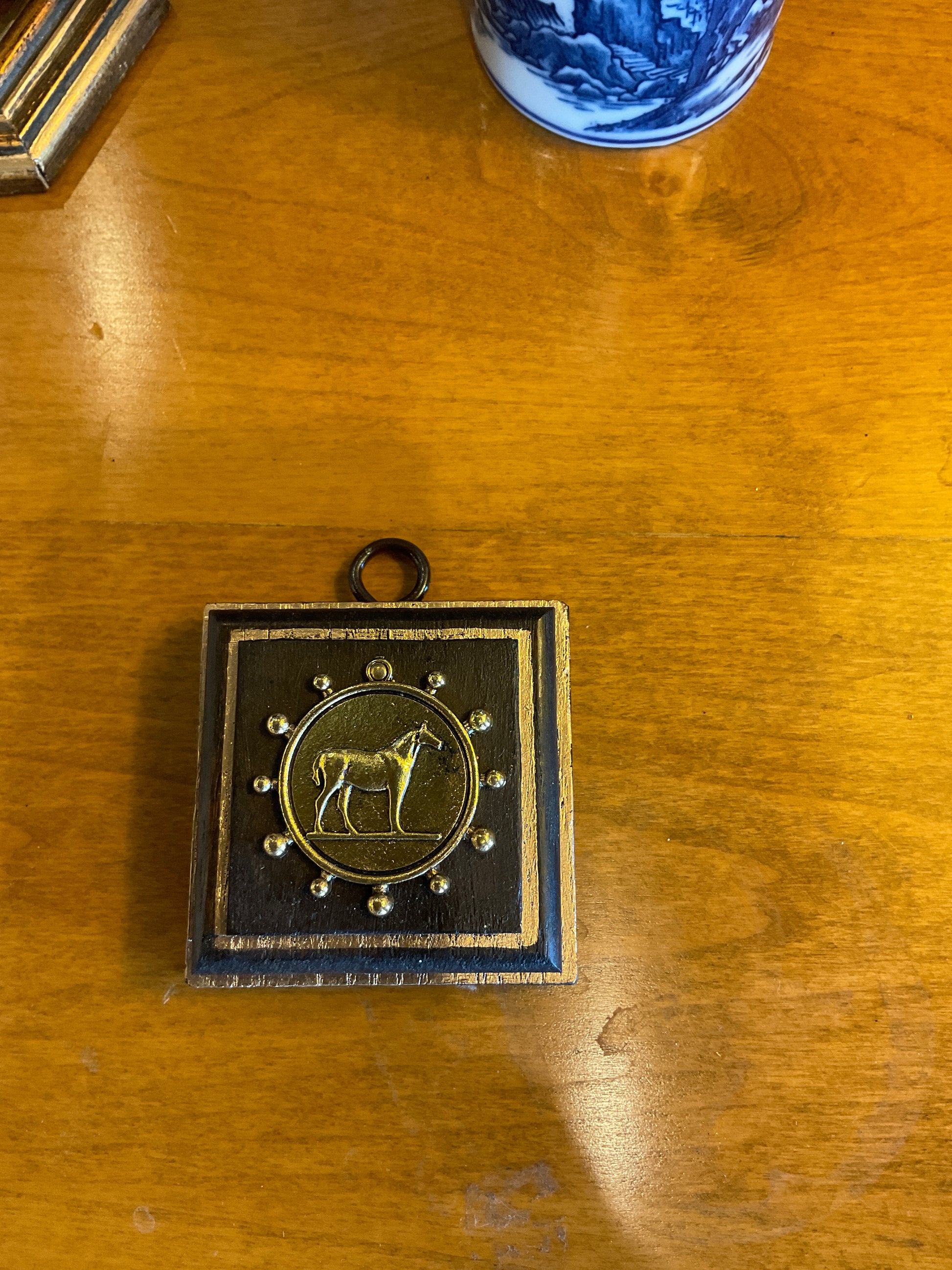 A square black and gold pendant or medallion featuring a central gold horse emblem, displayed on a warm wooden tabletop.